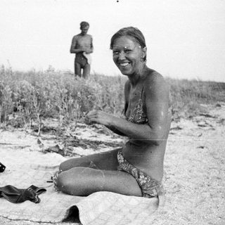 Woman sits on beach, smiling, with another person in background.
