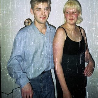 Here's a brief summary of the image: Young couple stands together, visibly aged photo with scratches.
