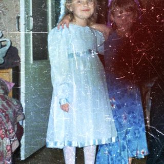Two young girls in dresses stand near a bedroom.