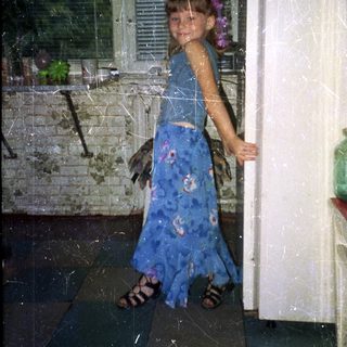 Girl in a blue dress stands near a refrigerator.