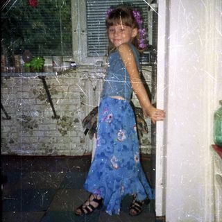 Girl in a blue dress stands near a refrigerator.