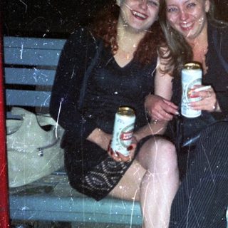Two women sit on a bench, holding drinks at night.