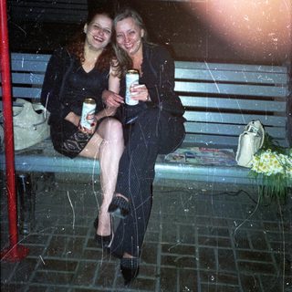 Two women sit on a bench, holding drinks at night.