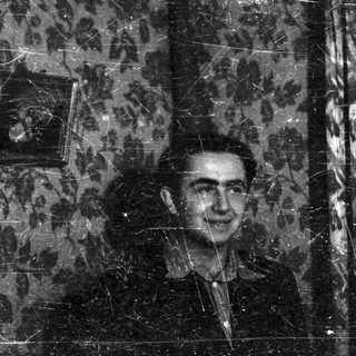 Black and white portrait of a man indoors with patterned wallpaper.