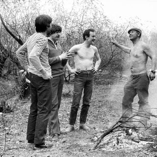 Men stand near a smoky fire in a wooded area.