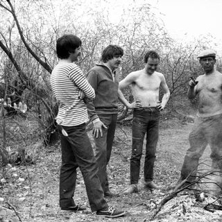 Four men standing near a fire in a wooded area.
