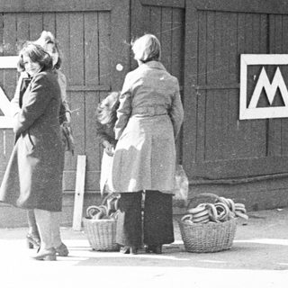 Two women stand near a fence with a sign.