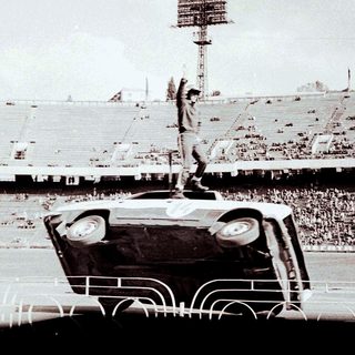 Man stands atop an overturned car in a stadium.