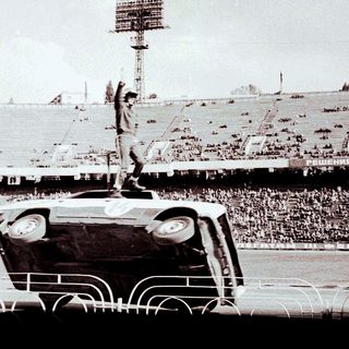 Man stands atop an overturned car in a stadium.