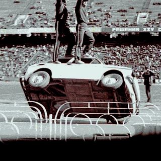 Man standing on car, raising arm in stadium setting.