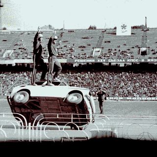 Man standing on car, raising arm in stadium setting.
