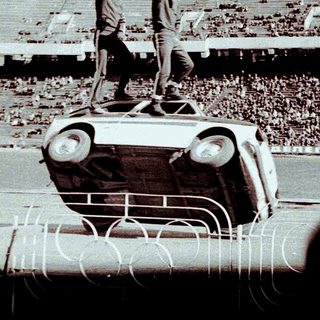 Man stands atop an overturned car in a stadium.