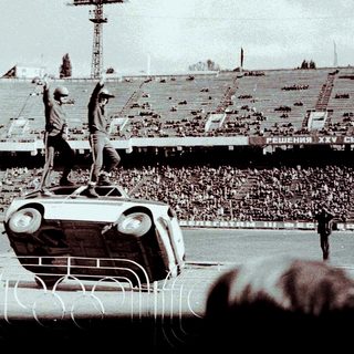 Man stands atop an overturned car in a stadium.