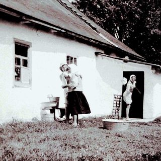 Here's a brief summary of the image: Two women stand outside a house in a rural setting.