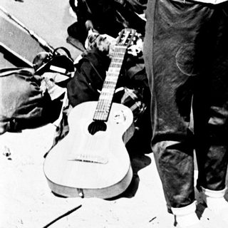 Here's a brief summary of the image: Couple with guitar, beer, and casual attire outdoors.