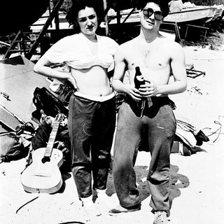 Here's a brief summary of the image: Couple with guitar, beer, and casual attire outdoors.