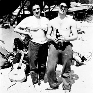 Here's a brief summary of the image: Couple with guitar, beer, and casual attire outdoors.