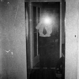 Here's a brief summary of the image: Figure reflected in a window, dark room, grainy image.