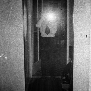 Here's a brief summary of the image: Figure reflected in a window, dark room, grainy image.