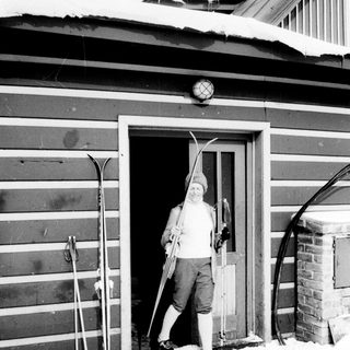 Man stands in doorway holding skis in snowy setting.