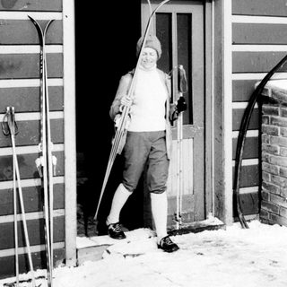 Man stands in doorway holding skis in snowy setting.