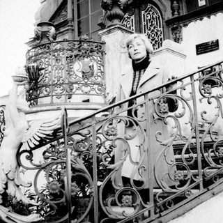 Woman stands on stairs with ornate railing and facade.