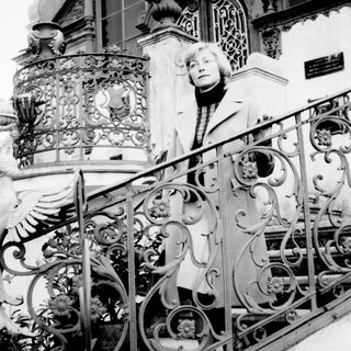 Woman stands on stairs with ornate railing and facade.