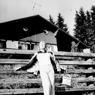 Woman stands by a wooden fence with a building behind.