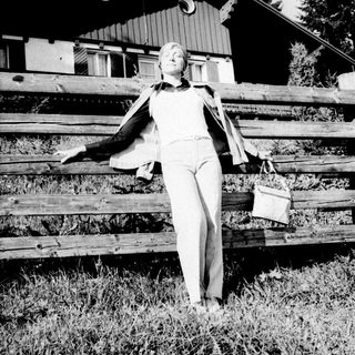 Woman stands by a wooden fence with a building behind.