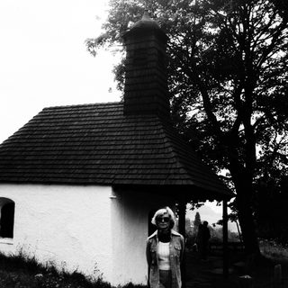 Woman stands near a small, rustic building in a landscape.