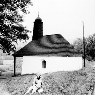 Woman sits near a small, rural chapel and landscape.