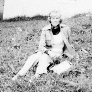 Woman sits near a small, rural chapel and landscape.