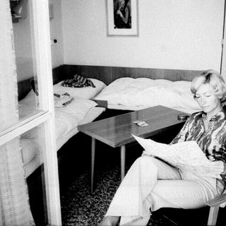Woman seated reading in a sparsely furnished bedroom.