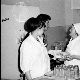 Two women in white coats talk in a kitchen setting.