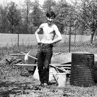 Man stands in a garden with barrels and chickens.