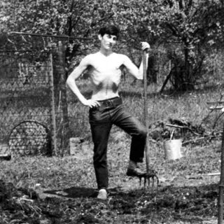 Man stands in a garden with tools and a fence.