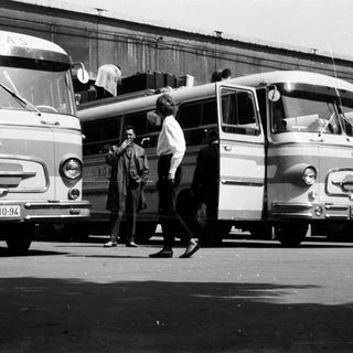 Black and white image of buses and people standing nearby.