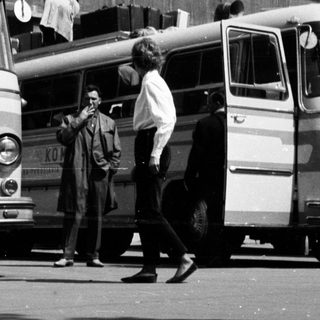Black and white image of buses and people standing nearby.