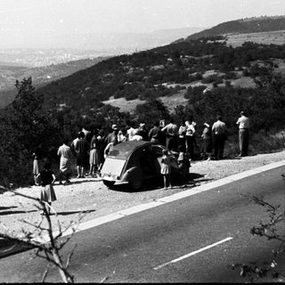 Group gathers near bus and car on a scenic road.