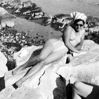 Woman sunbathing on rocks, reading a book by the sea.