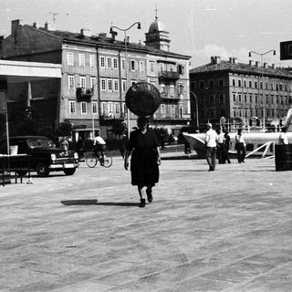 Woman carries basket on head in a town square.