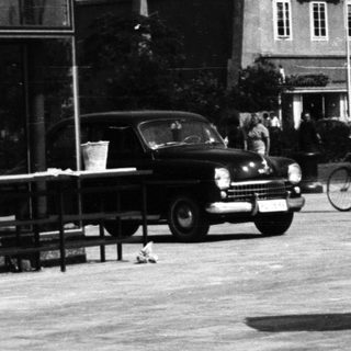 Woman carries basket on head in a town square.
