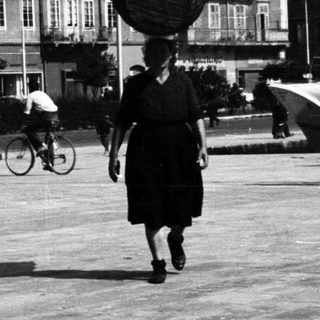 Woman carries basket on head in a town square.