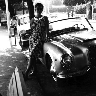 Woman stands beside vintage car on a public street.