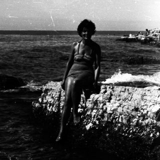 Woman sits on rocks overlooking a sea coastline.