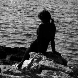Person sitting on rocks overlooking a sailboat on water.
