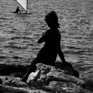 Child sits on rocks, watching a sailboat on water.