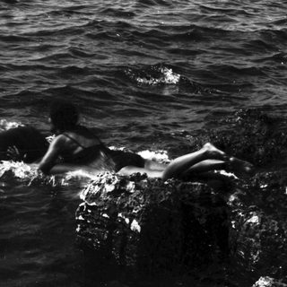 Person rests on rocks emerging from a dark sea.