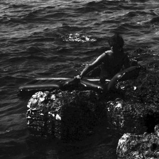 Black and white photo of figures on rocks in ocean.