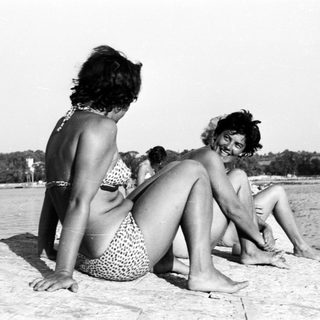 Here's a brief summary: Two women in swimwear relax by the water's edge.